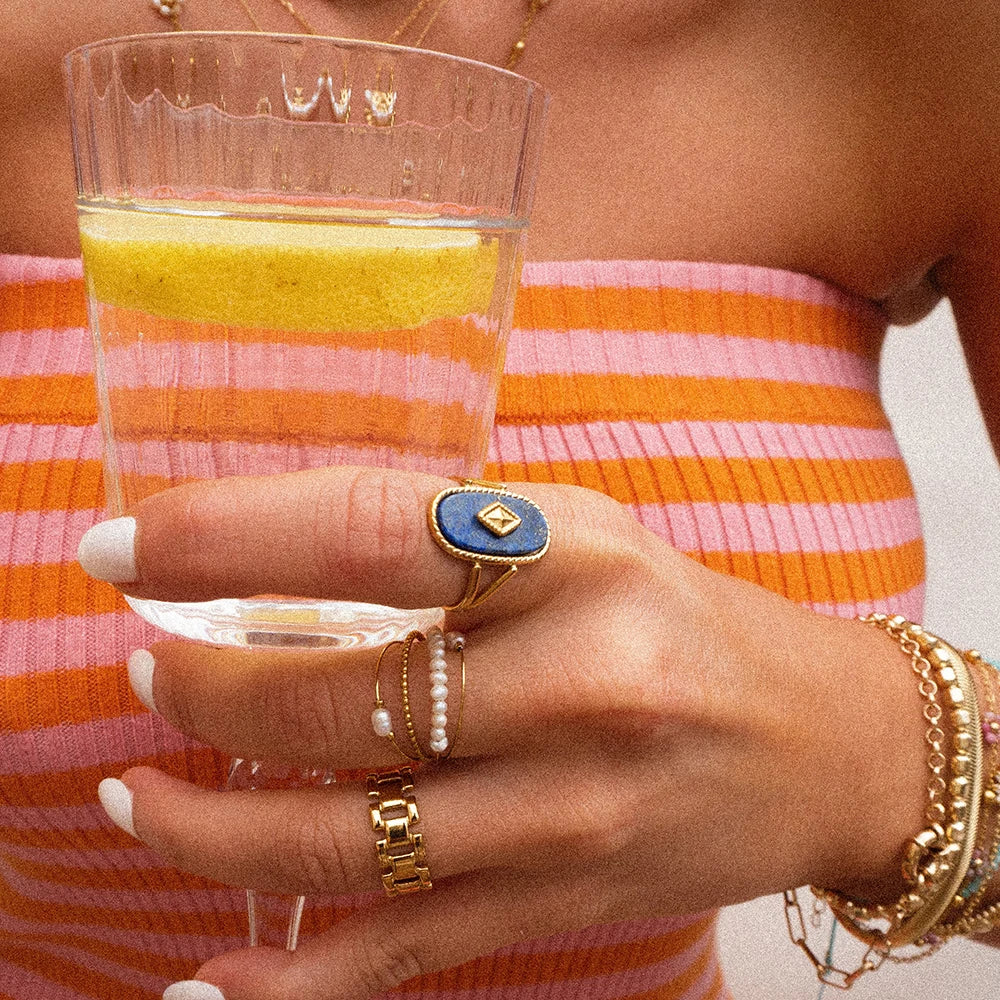Hand holding a glass of yellow liquid with jewelry, wearing an orange and pink striped garment.